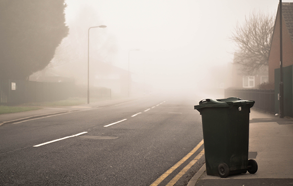 Image of a foggy, empty, residential street with a garbage can on the sidewalk